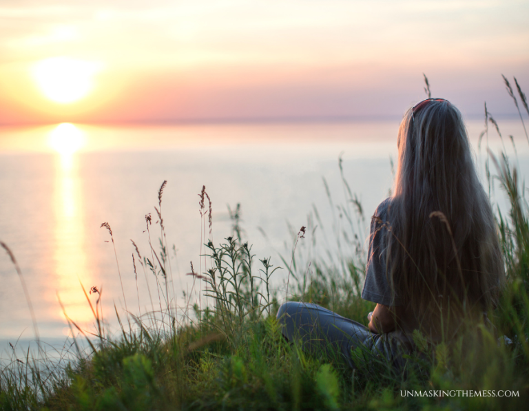 Christian woman sitting by a lake at sunrise reflecting on God’s Word.